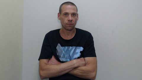 Phillip Humphrey has his arms folded and is looking at the camera in this police photo. He is wearing a dark T-shirt with a Nike logo and has closely shaven hair.