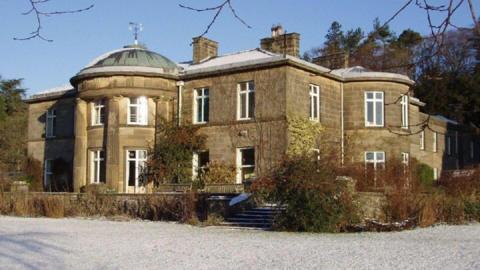 A large mansion-style building, pictured from the front. A frozen lawn covered in frost is in the foreground. A small set of steps lead up to the house itself, while there are also some shrubs around.  