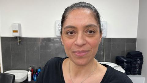 A woman with dark hair, tied back, smiles at the camera. She is wearing a black top and standing in front of a sink, what looks like shampoo bottles and towels.