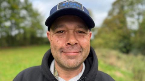 A man with a dark blue cap and black fleece standing in a green field