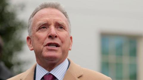 Steve Witkoff in striped shirt and red tie, wearing a brown coat