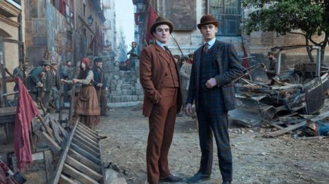 Young Sherlock stands with another man in an orange suit in a street made to look like a Victorian era London street. He wears a checked suit and a bowler hat