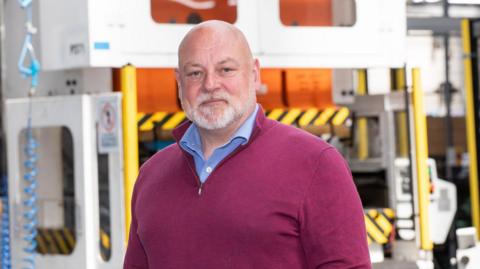 James Leng, who runs the manufacturing firm Pre-Met in Redditch, is stood on his factory floor wearing a red jumper and blue shirt. Behind him is a large yellow machine.