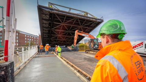 Drypool Bridge in Hull. Part of the structure is raised with workers standing beneath it. There is a mechanical digger in shot, along with a worker wearing an orange jacket and green safety helmet in the foreground.