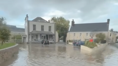 Flood water covers a road at a junction, with several cars parked up in front of buildings  showing the water reaches a few inches.