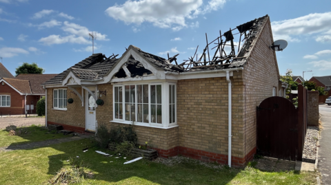 A view of the front of a bungalow in a residential street. Its damaged roof can be seen badly damaged and caved in.  