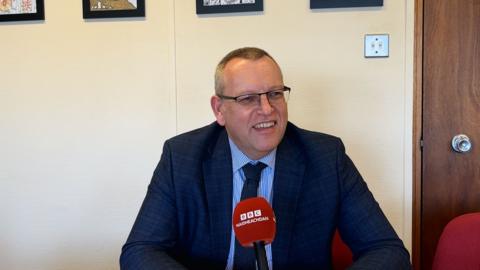 Dòmhnall MacLeòid, Comhairle nan Eilean Siar, na shuidhe san oifis aige agus gàire air aodann.