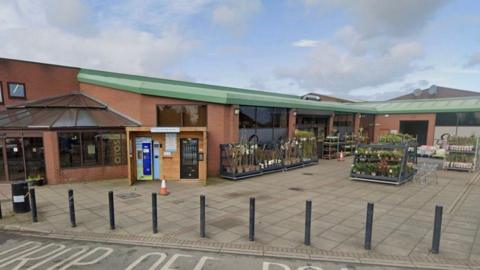 The exterior of a supermarket building with mainly a flat roof, with green panelling. There are bollards separating a drop off zone from the front of the store and there are rows of plants for sale outside.