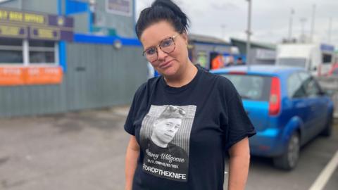 A woman with black hair tied in a pony tail. She is wearing glasses and a black T-shirt with the picture of a boy on the front with the words 'Harvey Willgoose #DroptheKnife'