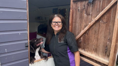 Karen is stood in the doorway of a shed with a dog leaning on a lower wooden door with two paws visible, looking at her. Karen is wearing a black and purple top. She has long hair and glasses.