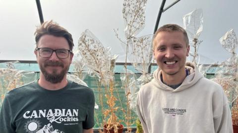 Two men are stood inside a plastic greenhouse in front of dying lettuce plants that have bags over the top of them. The man on the left is wearing glasses and a green t-shirt with white writing, he has brown hair and a beard. The man on the right has short blond hair and is wearing a grey hoodie with black writing. 