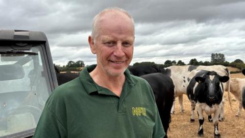 There is a man in a dark green polo shirt, with 'NFU' in yellow letters next to it. He has grey thin hair and is standing. To his left, there is his buggy, which is gret and has a window. To his right, there are cows.
