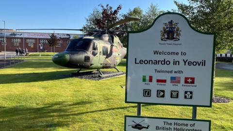 A sign reading "Leonardo in Yeovil" on a green field, with a camo-painted helicopter in the background.