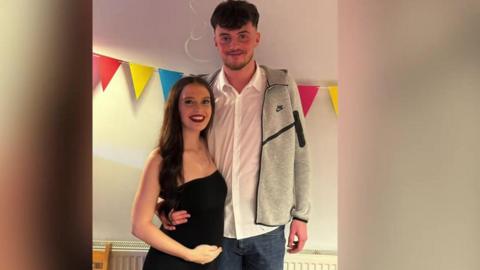 A young man and a young woman standing side-by-side with colourful bunting behind them. He is wearing a white, button-up shirt with a grey jumper over the top, and she is wearing a black dress and is holding her baby bump.