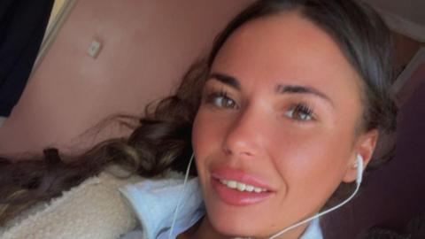 A woman takes a selfie of herself smiling. She has long black hair, with earphones in her ears and is wearing a white fluffy jacket over a blue top.