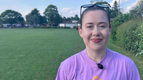 Woman with dark hair tied back and sunglasses on her head wears a mauve T-shirt. She is smiling at the camera and stands in front of a large park, with a hedge on the right of the photo.