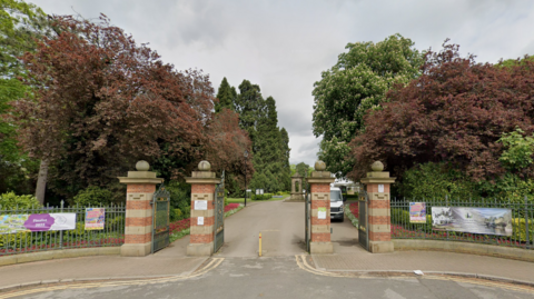 The entrance to South Park has four red brick columns with dark green metal gates between them. Lush green and brown trees can be seen in the park within.