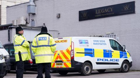 Two police officers wearing yellow jackets and black helmets stand outside a white bricked building. A crime scene investigation police van is parked underneath a black sign that reads Legacy Independent Funeral Directors