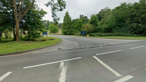 The Portsmouth Road entrance to Claremont Fan Court School is a large junction with chevrons and white road markings. In the school entrance there are two blue signs, with trees and fields.