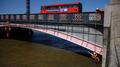 Lambeth Bridge in the sunshine with a red London bus going over it. An engineer is dangling off the bridge, carrying out repairs.