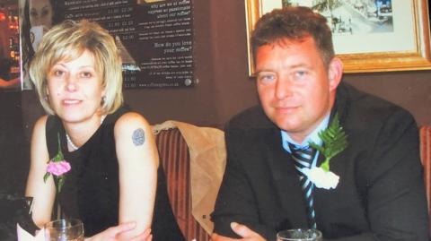 A woman on the left with blonde hair is next to a man wearing a dark jacket, shirt and tie. They are sitting down near a wall inside.