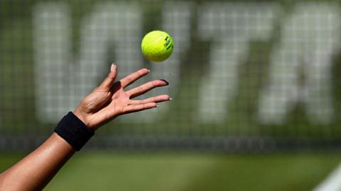 A female tennis player with long fingernails (only the hand shown) tosses a tennis ball in front of a net featuring WTA