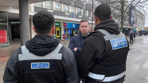 Gavin Callaghan, who has short brown hair and is wearing a navy jacket, with his arms folded as he listens to two community support officers. They are standing in Basildon town centre and dressed in black with their backs to the camera.