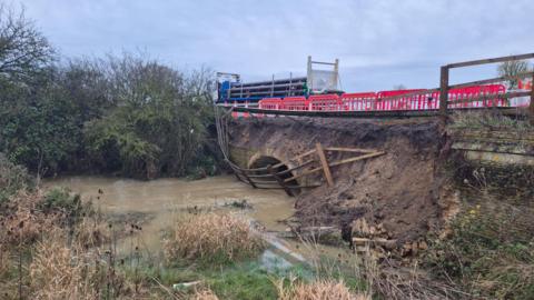 A damaged culvert under the A1 southbound over Billing Brook, Water Newton. It shows a damaged traffic barrier tipping over a muddy bank leading to the brick-built arched culvert opening. A wooden fence has tumbled down the bank and is tipped into the water, which is muddy with silt. There are shrubs to the left of the brook. A red crash barrier and red and white cones run along the edge of the road above the culvert.