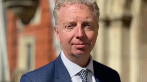 Alex Allinson is wearing a navy suit, light blue shirt and spotted tie. He's standing in front of a brick building and has short fair hair.