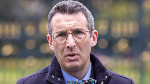 Andrew Muir is standing outside on front of large black gates. He wears glasses and is wearing a navy coat with a blue shirt and a bow tie.