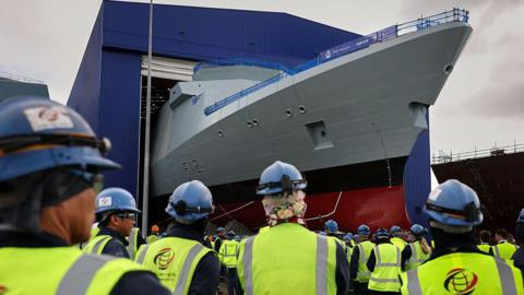 The Royal Navy frigate, HMS Venturer, being rolled out of the build hall at Babcock Rosyth. Dozens of workers in hi-vis jackets and blue helmets are watching with their backs to the camera 