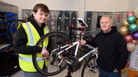 Two men standing in a bicycle repair shop. A white road bike is being held on a repair stand.