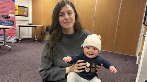 A young woman with long brunette hair wears a grey jumper. She has a smiling baby boy on her knee - he wears a white hat, navy jumper with bears on it and grey leggings.