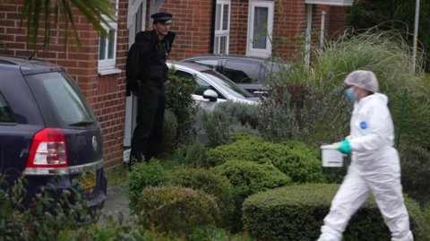 A police officer stands outside a red bricked, new-build house, while a forensic investigator walks up the driveway.