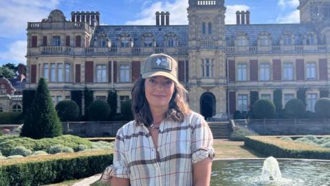 Lara Somerleyton stands in front of a fountain outside her home, Somerleyton Hall, a brick and cream stone Victorian-era mansion.
Lawns, pruned shrubbery and hedges are next to the fountain. A path and steps lead from the fountain to the entrance. 
