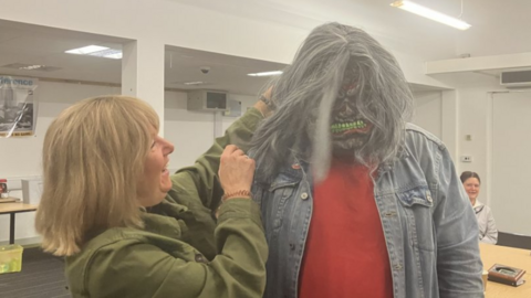 A woman with blonde hair and wearing a green jacket is putting a monster mask on a man who is standing next to her. The mask is grey and has grey long hair with green teeth. The man wearing the mask is wearing a denim jacket and a red sweatshirt.