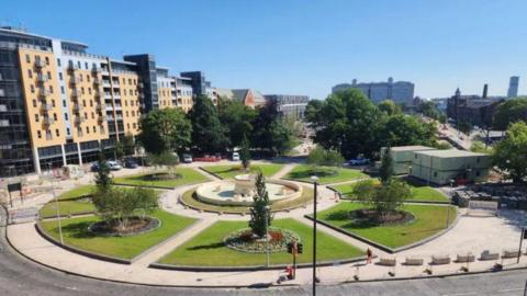 A view of the Rose Bowl and fountain in Hull as construction work takes place in Queen's Gardens, Hull. The cream coloured fountain stands in the centre of the image surrounded by a circular path and large, raised lawns and flower beds with trees. Construction fencing and vehicles are visible in the background.
