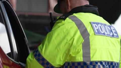 A male police officer wearing a high-viz yellow jacket with police written in white letters on a blue rectangle on his back, standing beside a red car with an open window