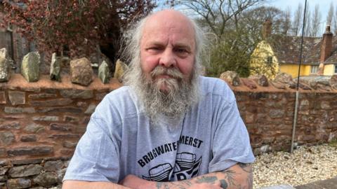 A man in his 60s sits with his arms folded and resting on a table outside. He has a mostly bald head with wispy grey hair and a long beard. He is wearing light blue T-shirt with a motif on it and text that reads "Bridgwater, Somerset". Behind him is a brick wall, a large yellow house and trees.
