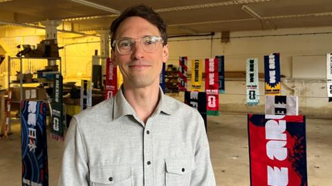 A picture of Tom Pitts, who has dark brown hair and glasses, standing in front of a display of his World Cup scarves.