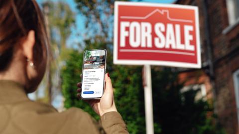 Woman looks at smart phone screen about a property with a for sale sign in the background.