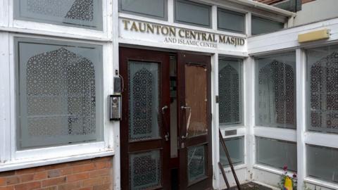The outside of a mosque. There is a sign above the door reading "Taunton central masjid and Islamic centre" in capital letters. Below the sign there are dark brown double doors with patterned glass panels. One of the doors is boarded up as the glass has been smashed.