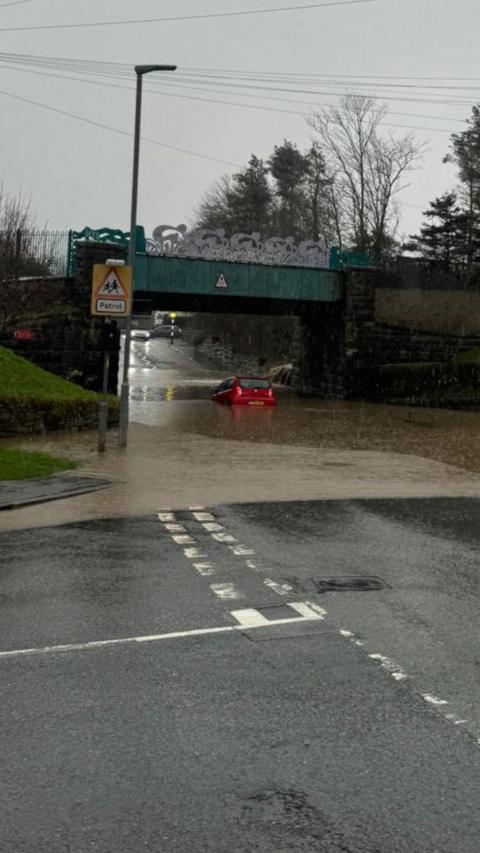 A junction on a rainy day. Flood water can be seen pooling under a bridge and there is a small red car in the middle of it.