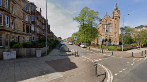 A baronial sandstone building and blond and red sandstone tenement flats stand on either side of Dixon Avenue, and the junction with Cathcart Road. Cars are parked on the roadside beyond a 20mph sign and a bike lock-up box.