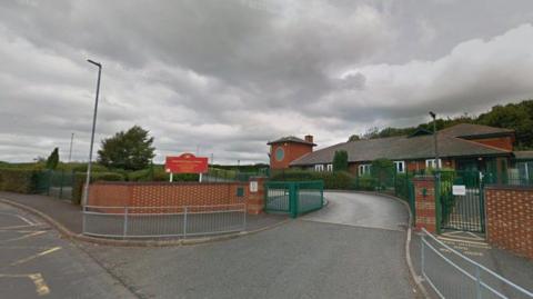 The entrance to the car park of Goldenhill Primary Acdemy, with a red sign indicating the name of the school. There is a single-storey building in the background with a number of bushes and trees around.
