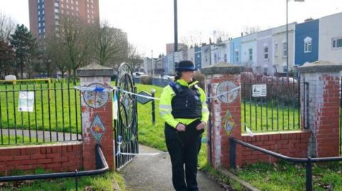 A police woman stands at an entrance to Rawnsley Park near to the scene in the St Philips area of Bristol where a 16-year-old boy died after being stabbed. There is police tape across the gate and in the background coloured houses can be seen and a tall tower block.