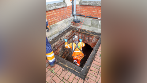 A man in a hi-vis jacket and a hardhat descends through a hole in a brickwork floor.