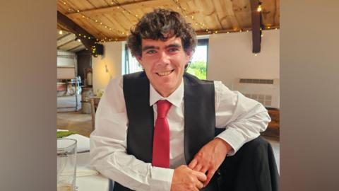 A man wearing a white shirt, dark grey waistcoat and red tie. He has dark hair and is smiling. 