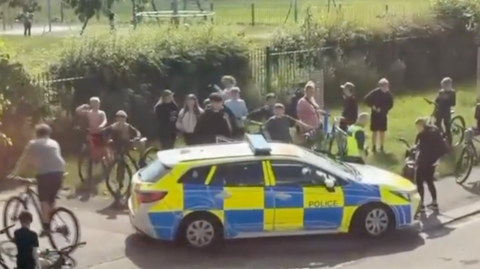 A group of about 20 mainly young people, most on pedal bicycles, are milling around a blue and yellow police car which has stopped near a park. Some are riding their bikes, while others have stopped and are standing with their bikes.