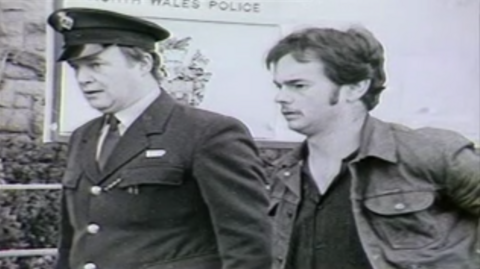 Black-and-white picture of a prisoner (on the right) being escorted by a prison officer. The prisoner is wearing a denim jacket and has a moustache.